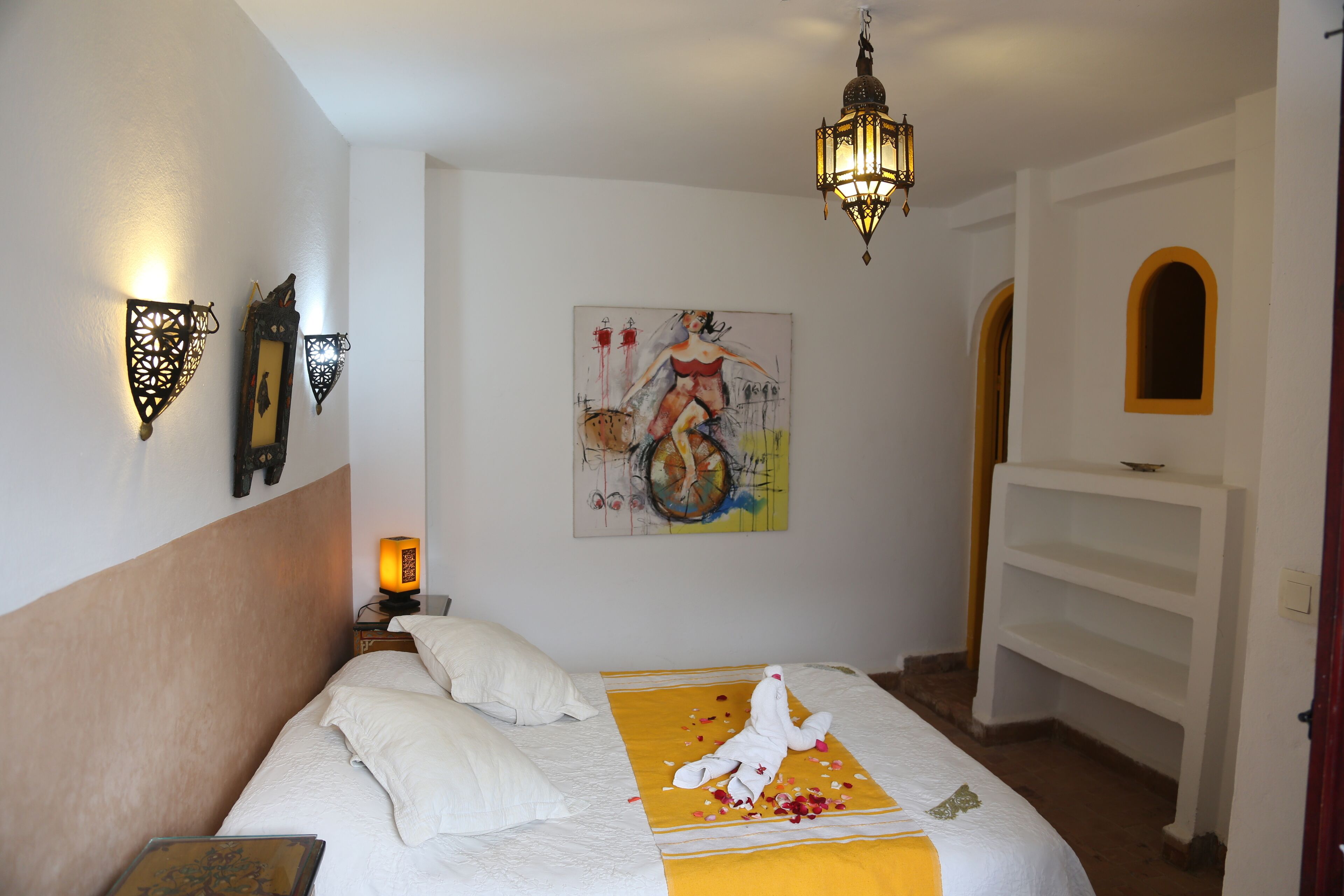 A hotel room with a bed, a painting on the wall, a nightstand, and a decorative wall light.