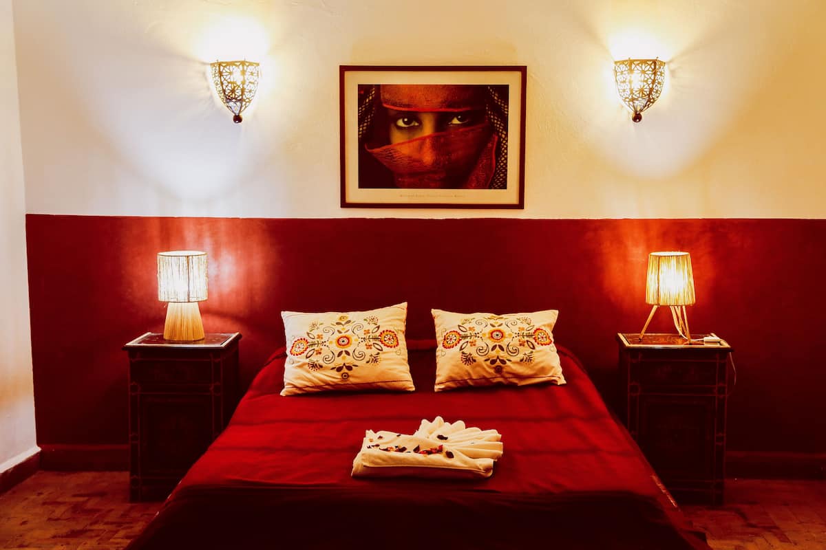 A hotel room with a red bedspread, two decorative pillows, a framed picture on the wall, and two bedside lamps.