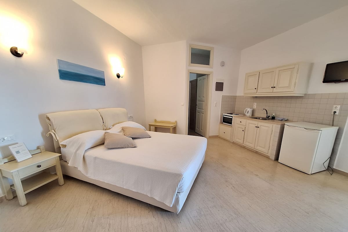 A bedroom with a bed, bedside tables, a nightstand, a wall-mounted TV, and a kitchen area with cabinets and a refrigerator.