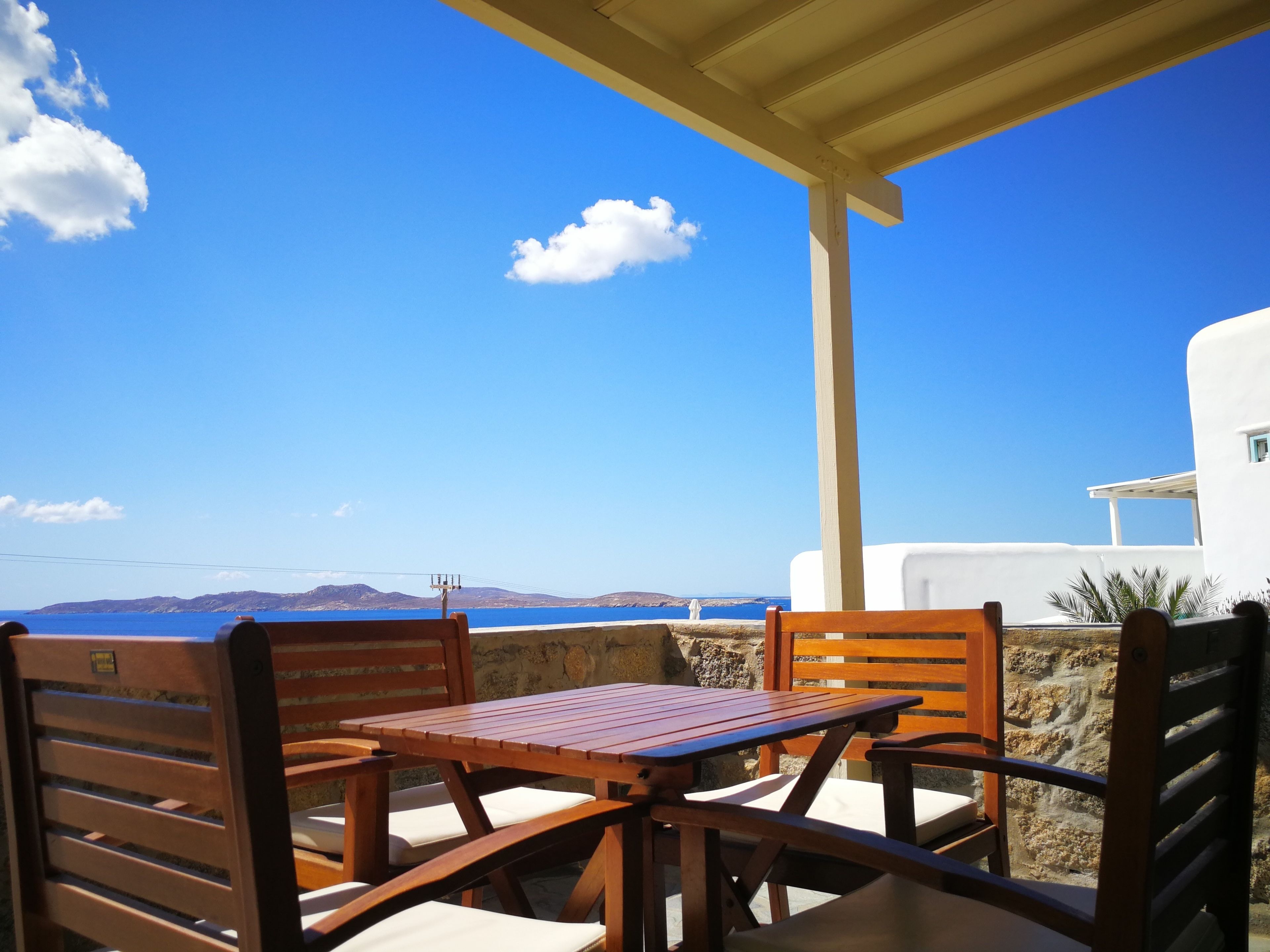 A wooden table and chairs on a balcony with a clear view of the sea and distant hills.