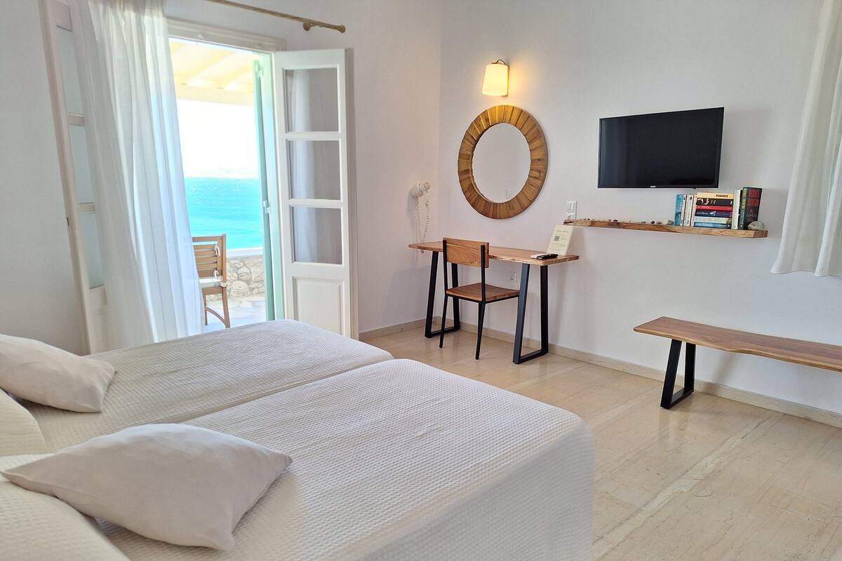 A hotel room with a bed, a desk, a chair, a TV, and a view of the sea.