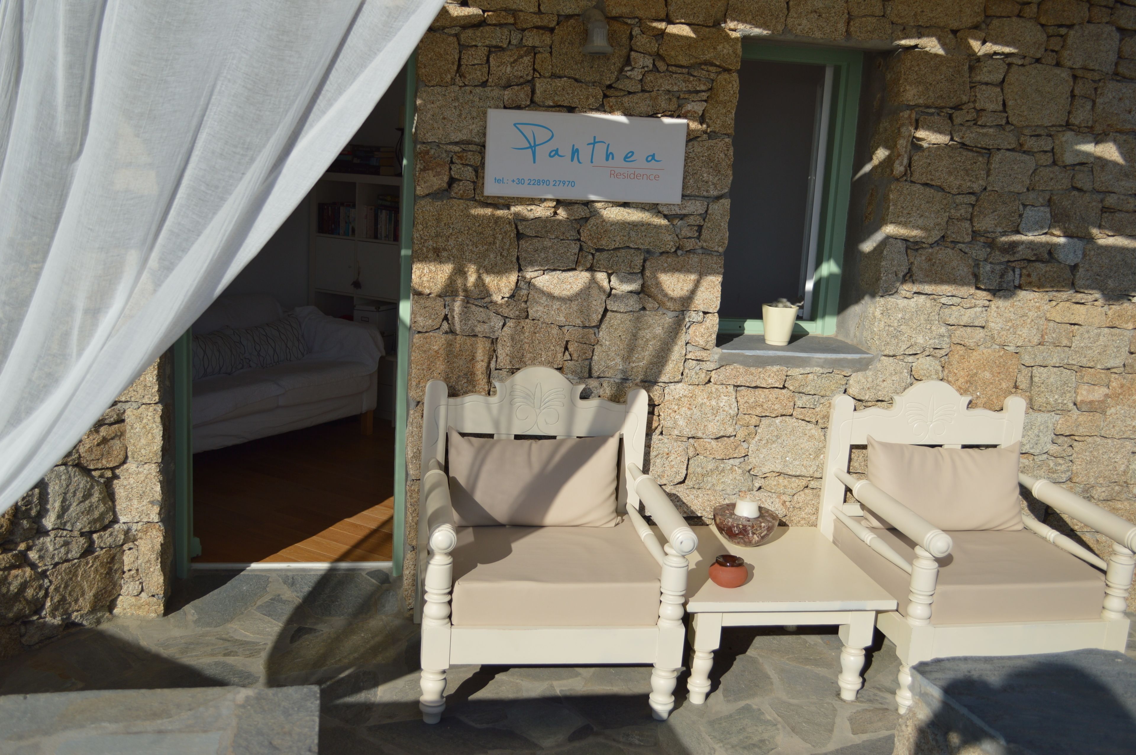 Un edificio in pietra con un cartello che riporta la scritta "Panthea Residence". Sono presenti due sedie bianche con cuscini e un tavolino con una ciotola e una candela.