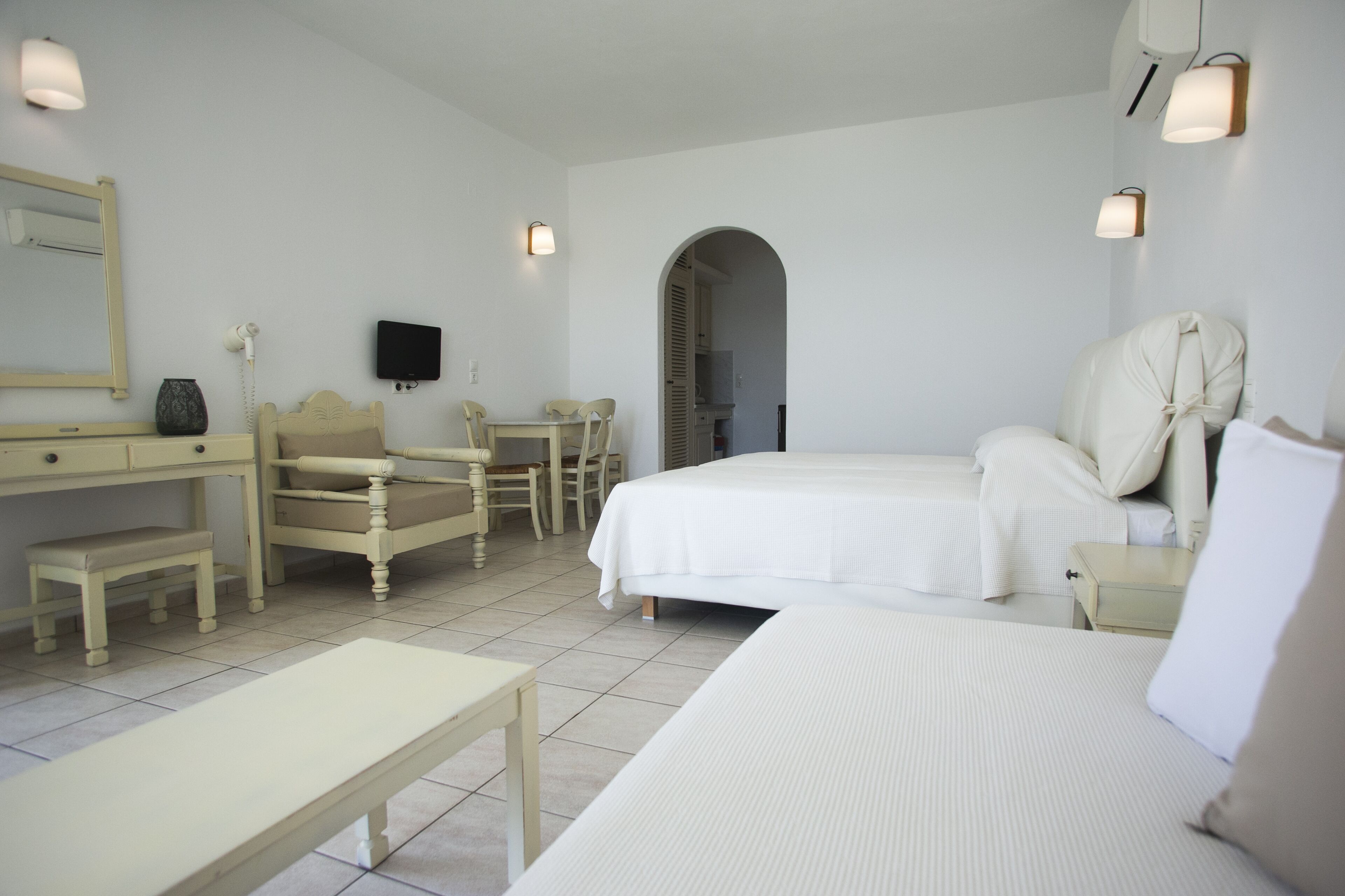 A hotel room with two beds, a desk, a chair, a small table, a TV, and a bathroom visible through an archway.