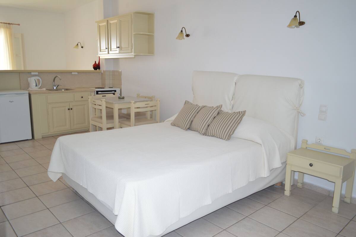 A bedroom with a bed, bedside table, and a small desk with a chair. There is a kitchenette with a refrigerator, stove, and sink.