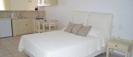 A bedroom with a bed, bedside table, and a small desk with a chair. There is a kitchenette with a refrigerator, stove, and sink.