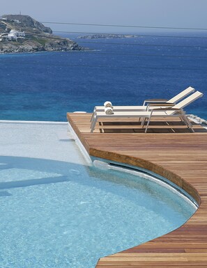 A wooden deck with lounge chairs and a pool, overlooking a clear blue sea and rocky coastline. - Panthea Residence (Mykonos)
