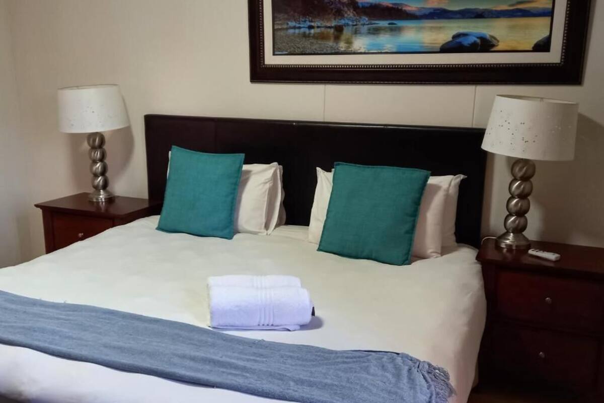 A neatly made bed with a blue blanket, two green pillows, and a white towel. There are two bedside tables with lamps, and a painting above the bed.