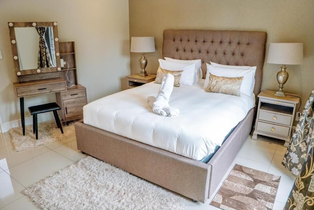 A bedroom with a large bed, two bedside tables, a dresser, a mirror with lights, and a fluffy rug.