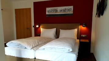 Un lit double avec du linge de lit blanc, une porte en bois, un mur rouge et une œuvre d’art encadrée au-dessus du lit.