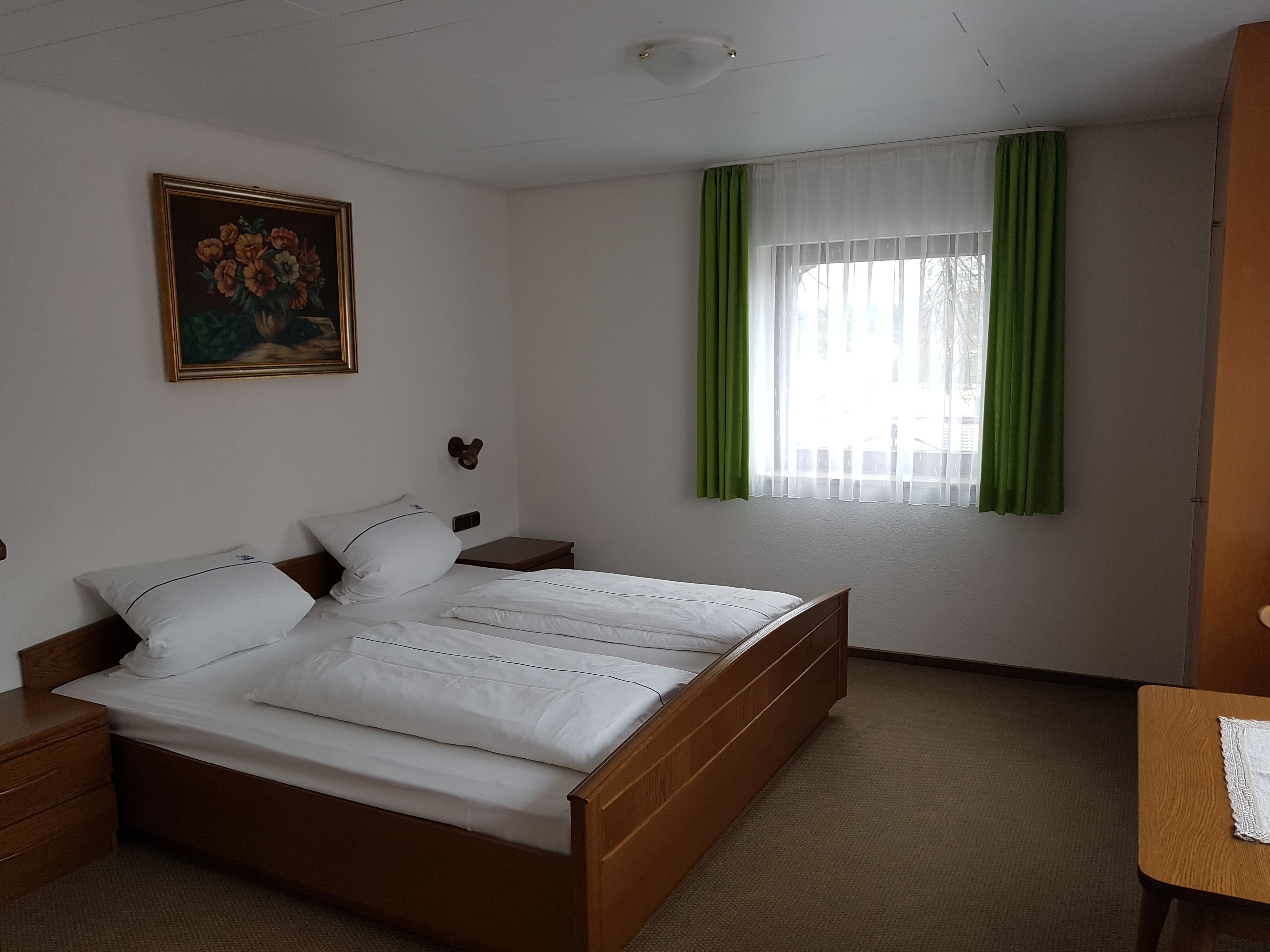 Ein Schlafzimmer mit einem hölzernen Bett, weißer Bettwäsche, einem Nachttisch, einer Wandlampe, einem Gemälde und einem Fenster mit grünen Vorhängen.