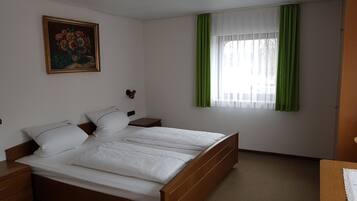 Ein Schlafzimmer mit einem hölzernen Bett, weißer Bettwäsche, einem Nachttisch, einer Wandlampe, einem Gemälde und einem Fenster mit grünen Vorhängen.
