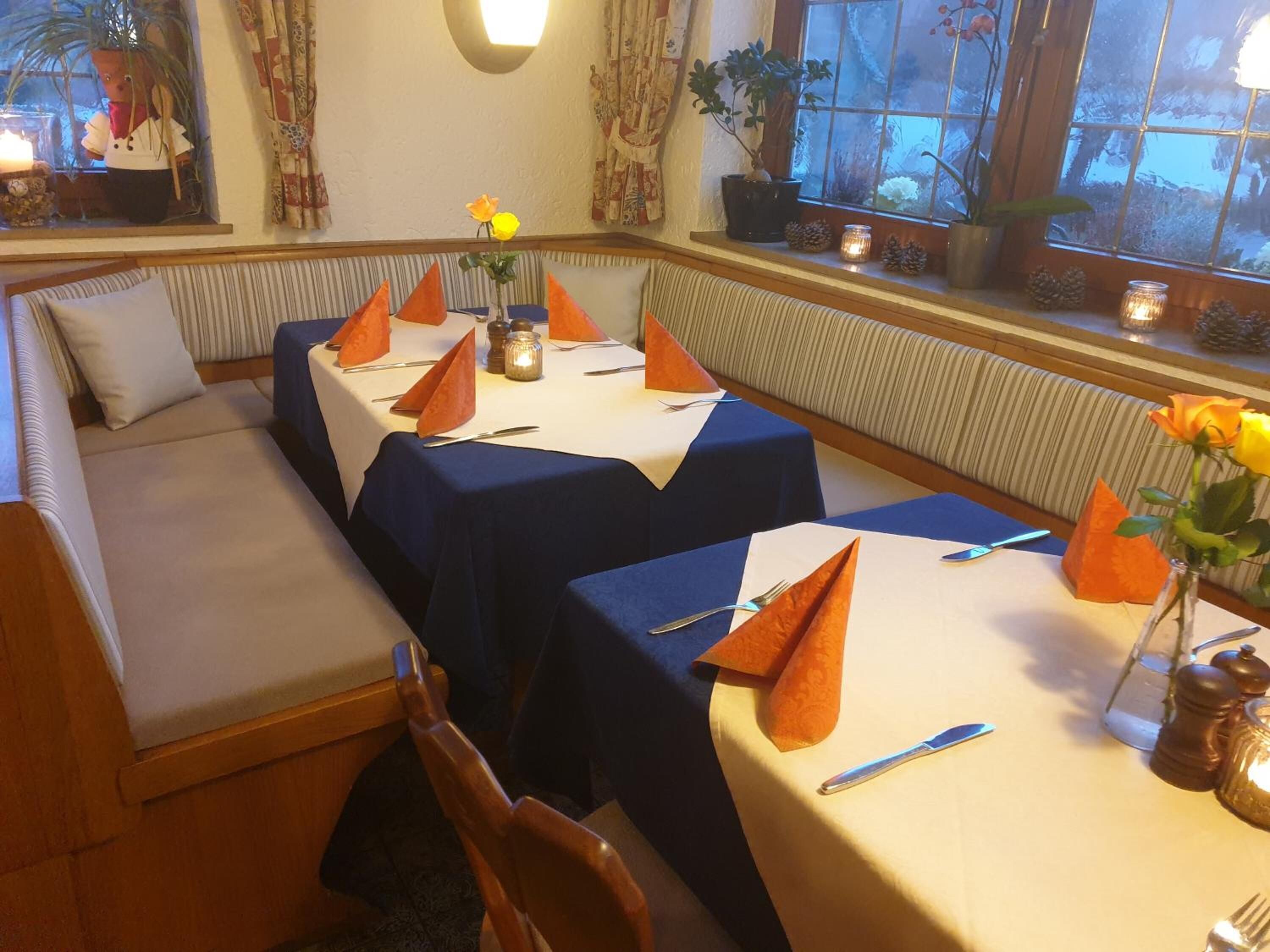 Un comedor con mesas dispuestas para cuatro, cada una con servilletas anaranjadas, un florero con una flor amarilla y una vela.