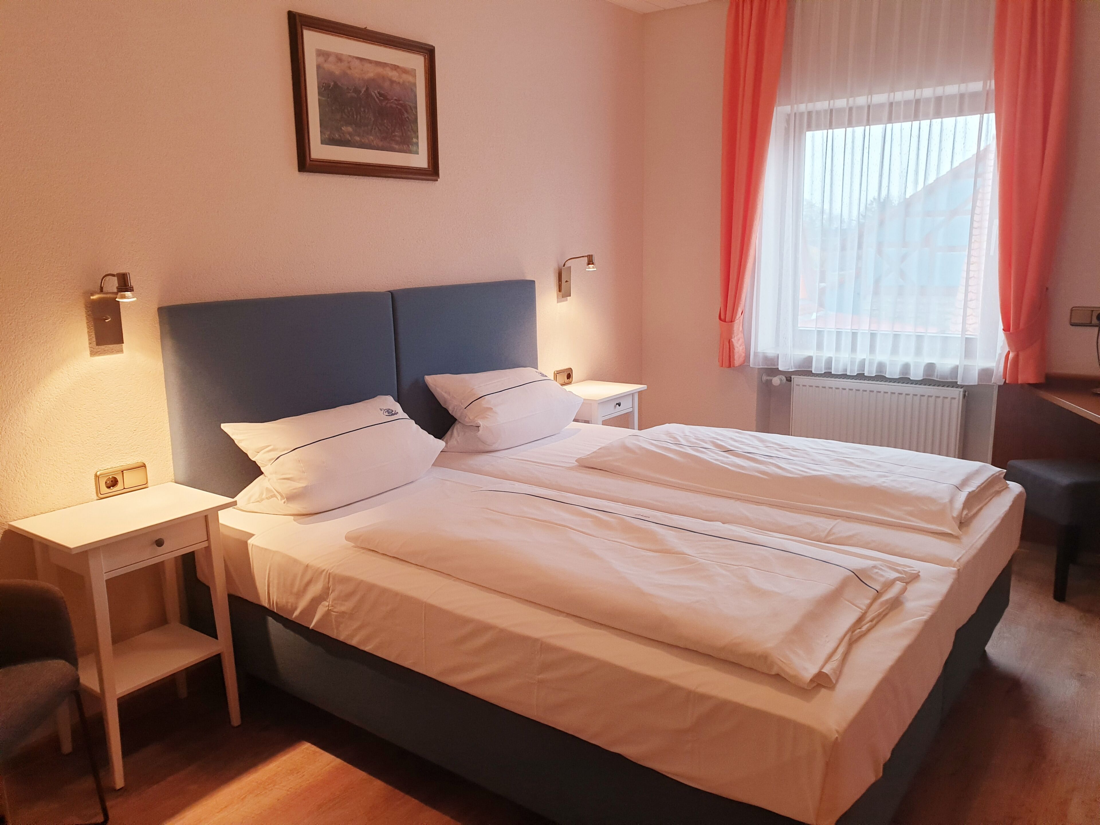 A double bed with white linens, a nightstand with a lamp, a small table, and a chair.