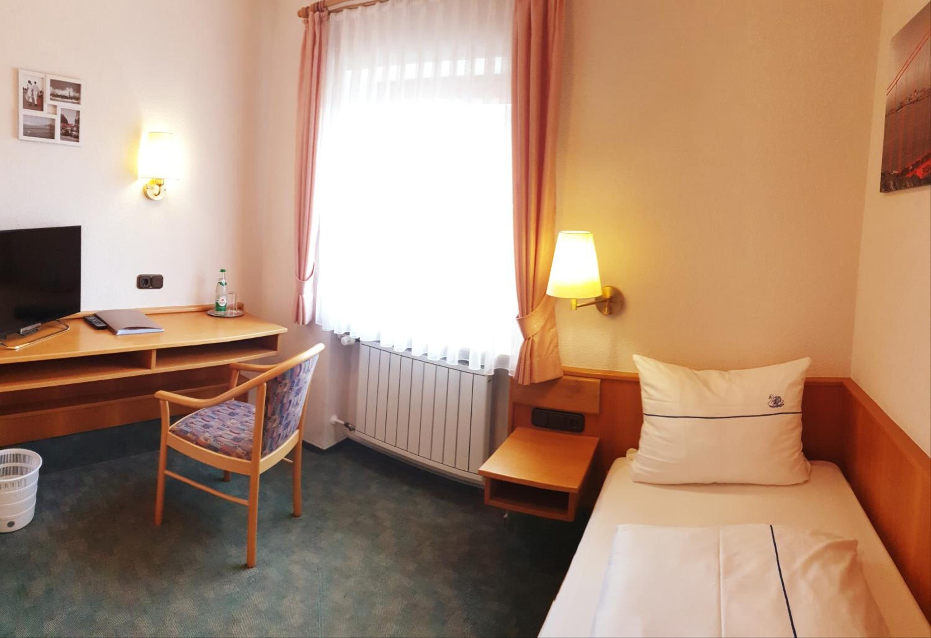 Habitación de hotel con cama, escritorio con computadora, silla, radiador, ventana con cortinas y lámpara de pared.