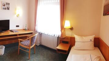 Habitación de hotel con cama, escritorio con computadora, silla, radiador, ventana con cortinas y lámpara de pared.