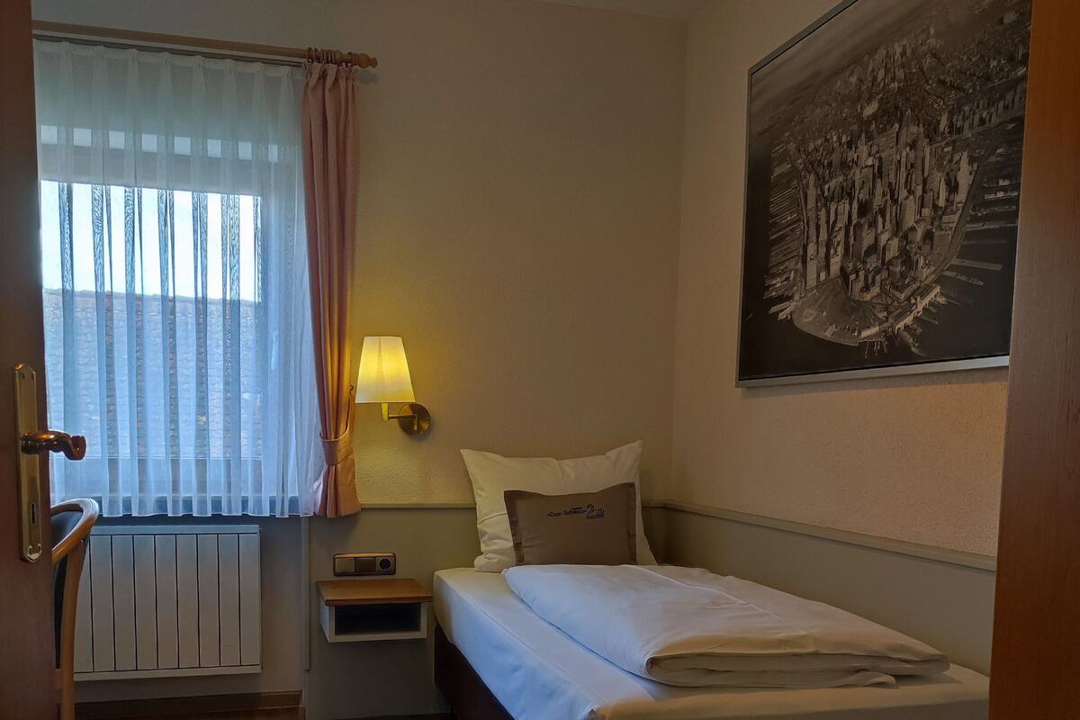 A hotel room with a bed, a nightstand, a lamp, a window with curtains, a radiator, and a painting on the wall.