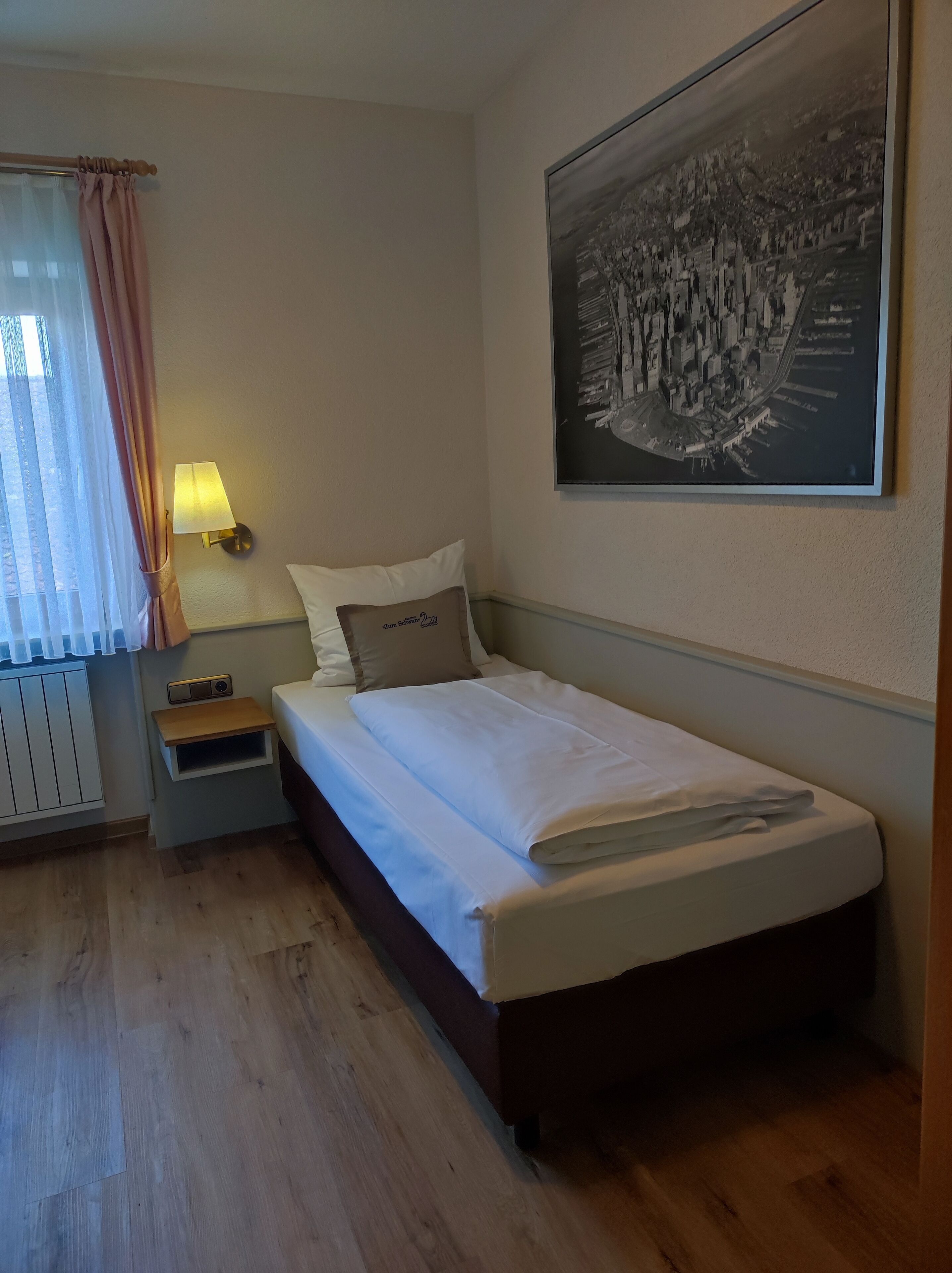 A neatly made bed with white linens, a bedside lamp, a small bedside table, and a wall-mounted picture of a cityscape.