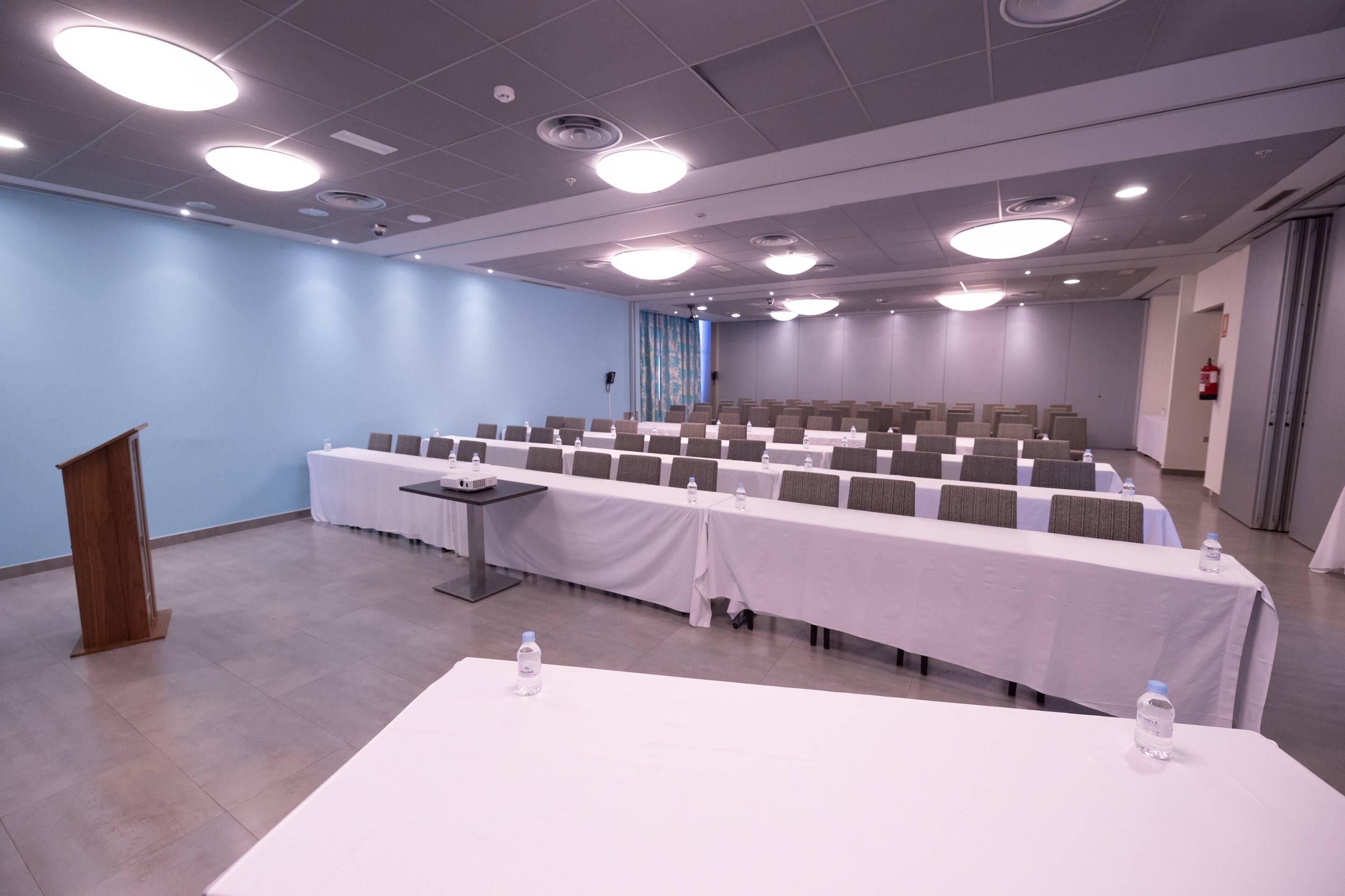 Un salón de conferencias con una larga mesa, filas de sillas, un atril y luces en el techo.