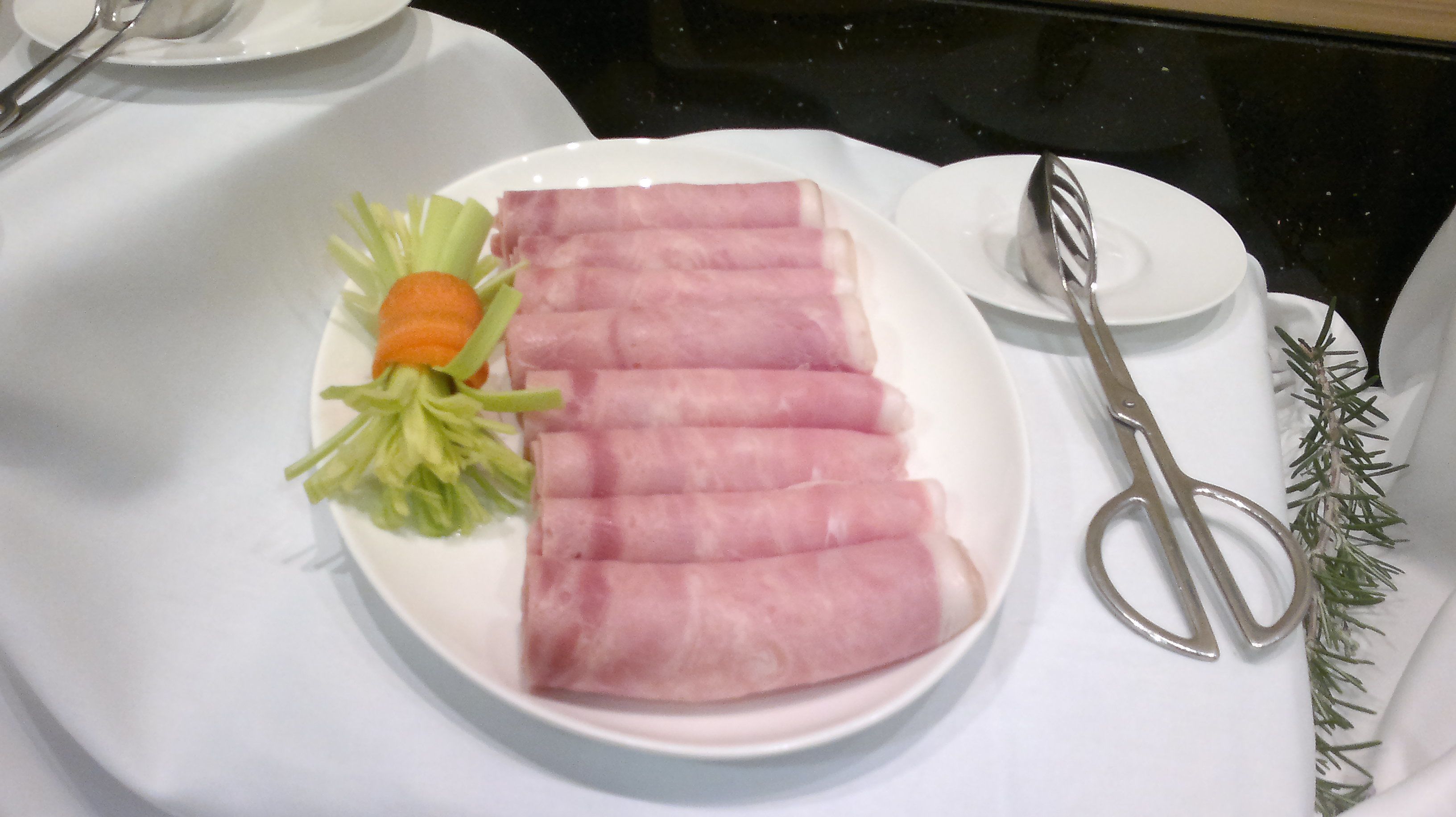 A plate with sliced ham, a carrot, and some greens on a white tablecloth.
