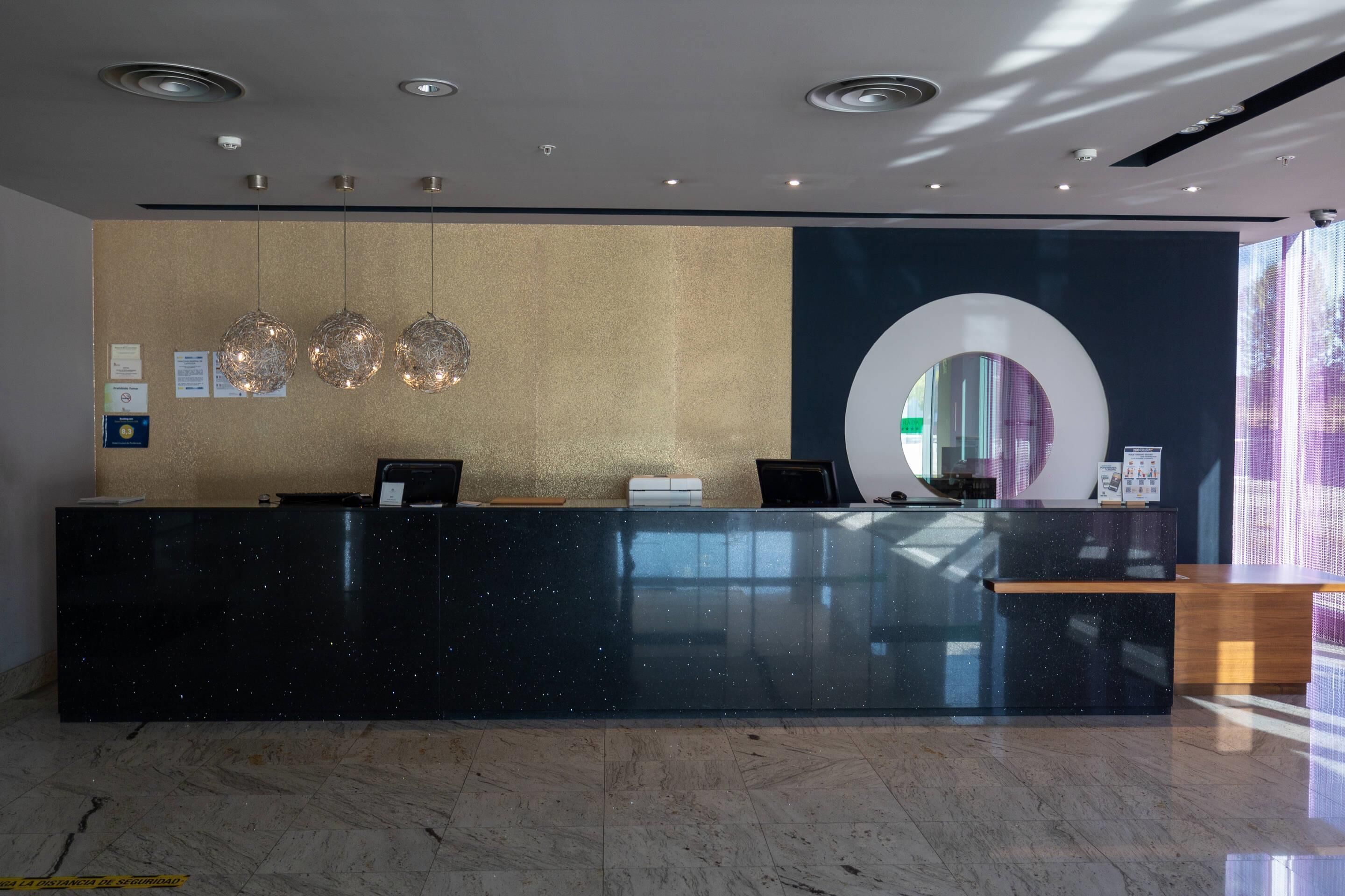 Zona de recepción de un hotel con un mostrador de mármol negro, tres lámparas colgantes esféricas y un espejo circular grande en la pared.