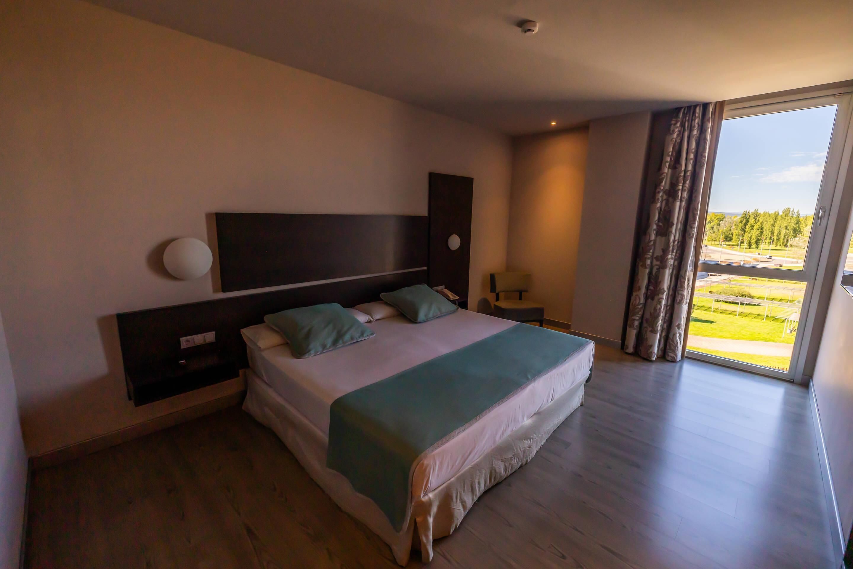 A bedroom with a large bed, a nightstand, a lamp, and a view of the outdoors through a window.