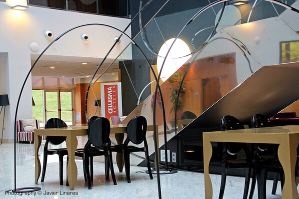 Un moderno vestíbulo de hotel con una escalera central, un mostrador de recepción y un área de descanso con mesas y sillas.