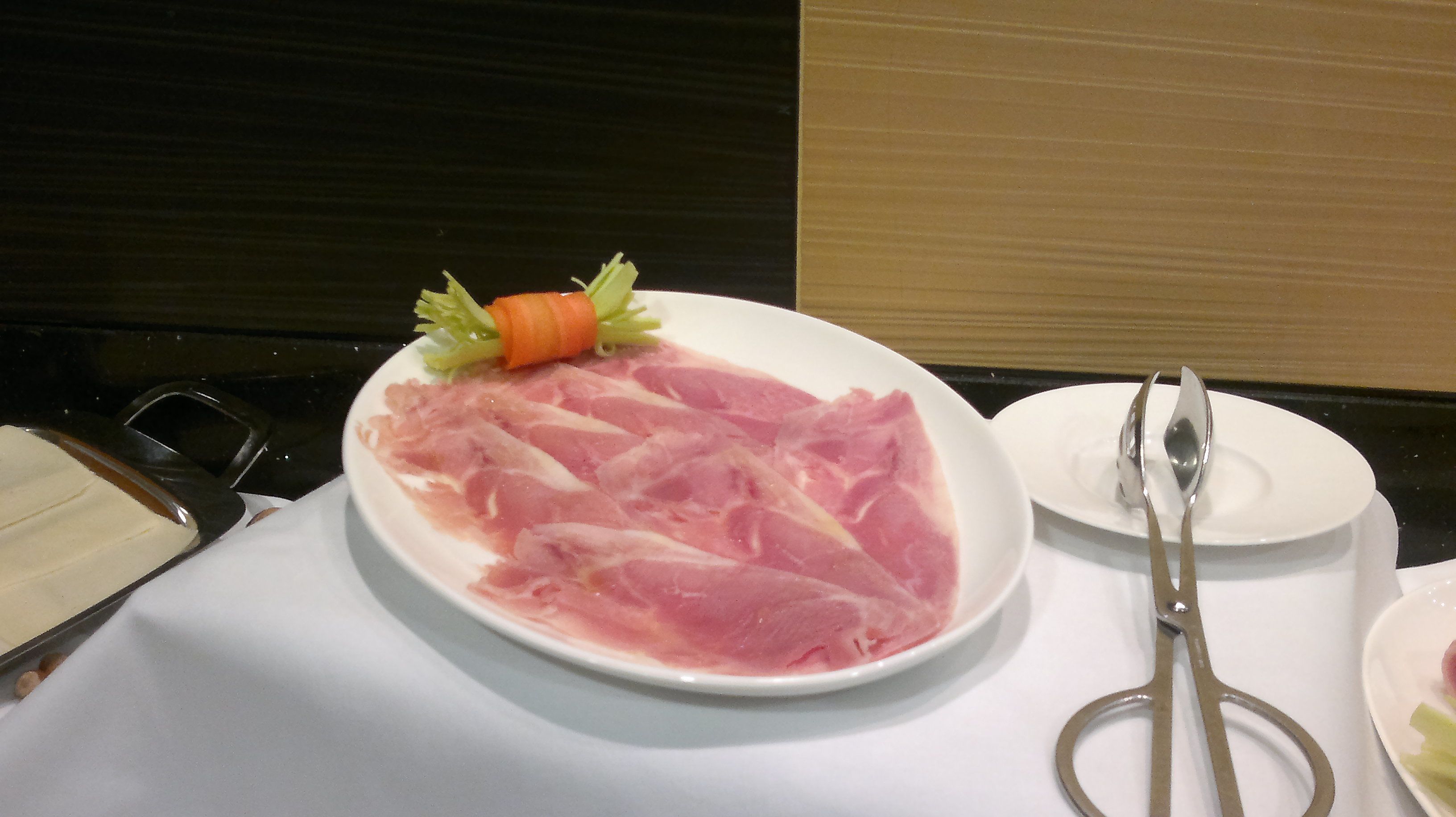 A plate of raw meat with a carrot garnish on a white plate, accompanied by a pair of scissors and a spoon on a table.