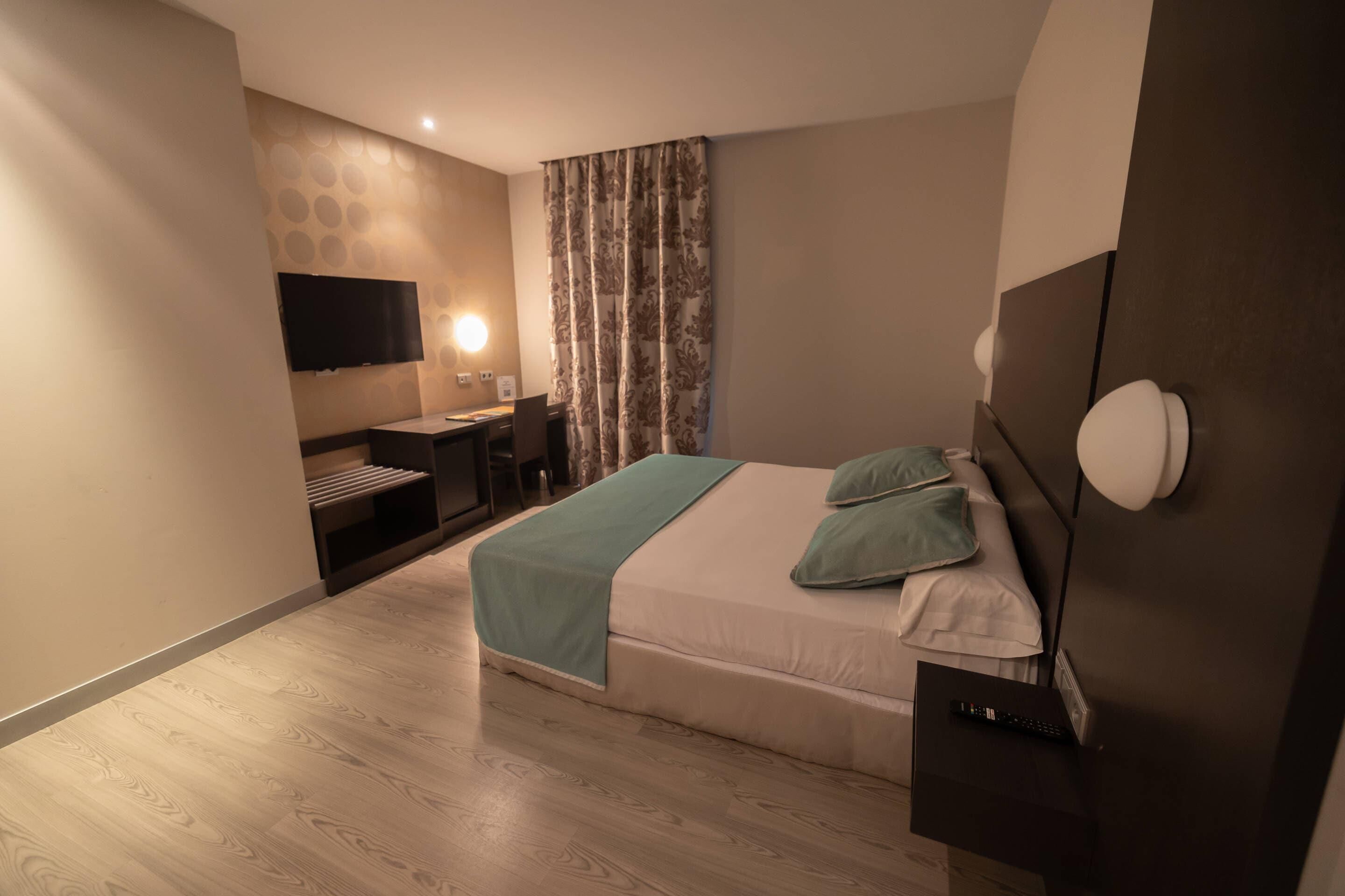 A modern bedroom with a bed, a desk, a chair, a flat-screen TV, and a lamp.