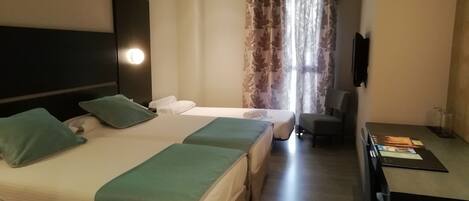 Habitación de hotel con dos camas, cada una con almohada verde y ropa de cama blanca. Hay un escritorio con una computadora portátil y una silla, y una ventana con cortinas.