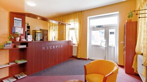 Un área de recepción con un mostrador de madera identificado con la leyenda "RECEPCIE", una silla amarilla y una ventana con cortinas.