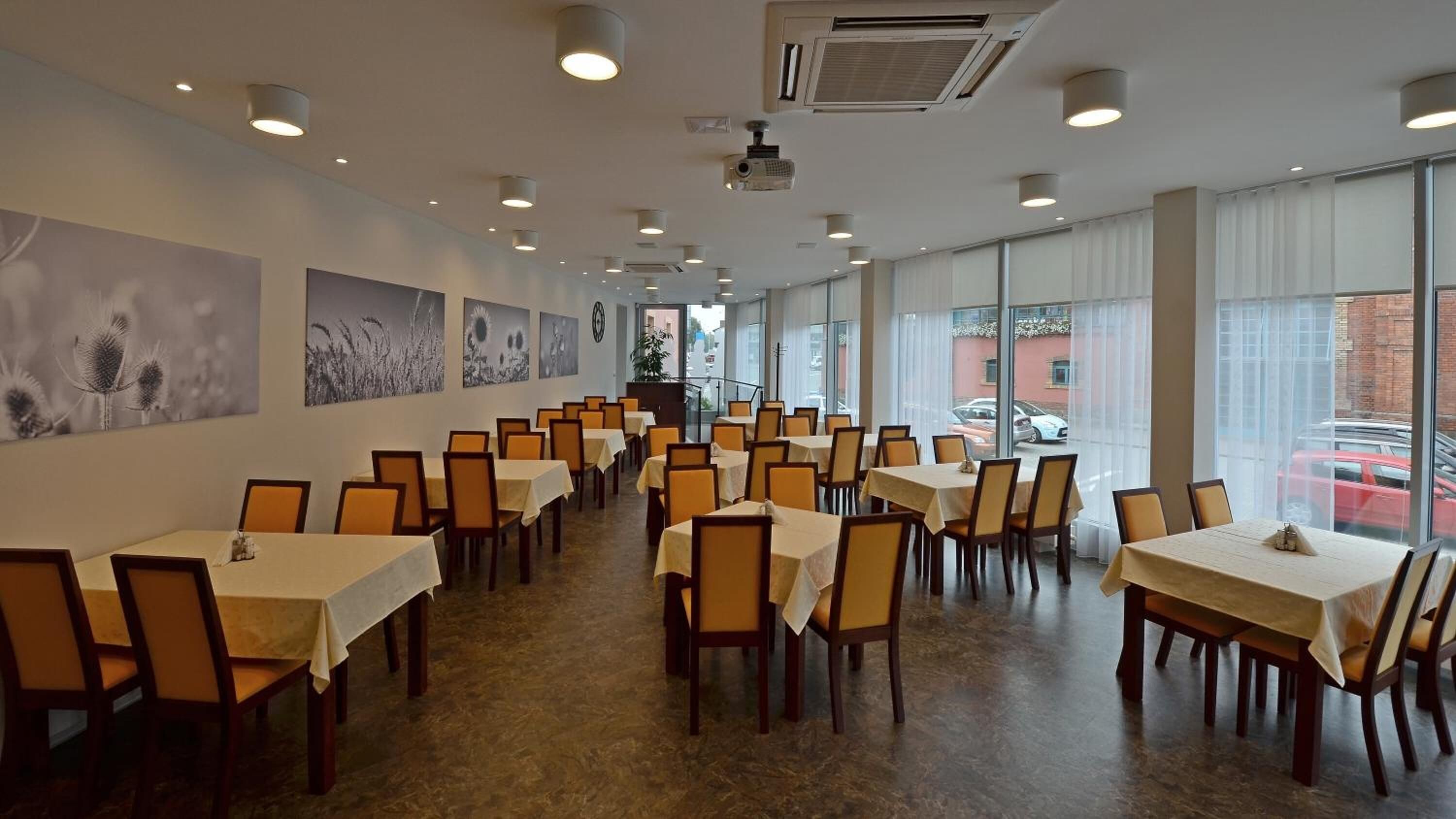 A spacious dining area with multiple tables and chairs, large windows with a view of the street, and wall-mounted artwork.