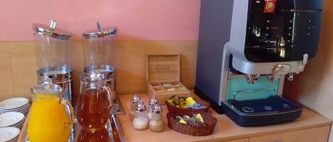 Una barra de desayuno con máquina de café, dispensadores de té y una cesta de frutas.