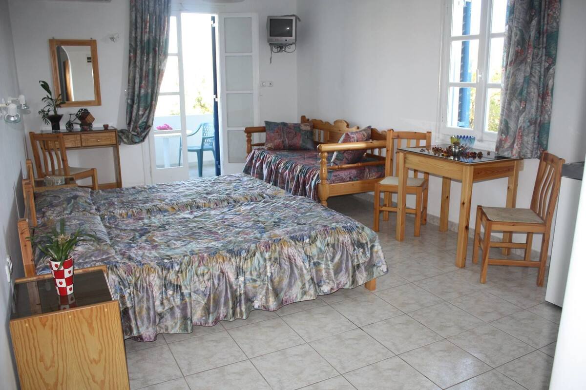 A bedroom with a bed, a desk, and a chair. There is a window with curtains, a mirror, and a television on the wall.