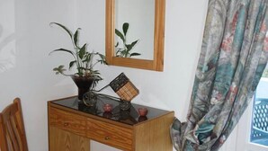 Une petite pièce avec un bureau en bois, une chaise, une plante en pot, un miroir et une fenêtre avec un rideau.