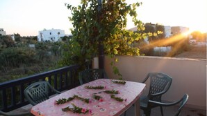 Une table recouverte d’une nappe à motifs floraux, entourée de chaises, sur un balcon extérieur offrant une vue sur des bâtiments et de la verdure.