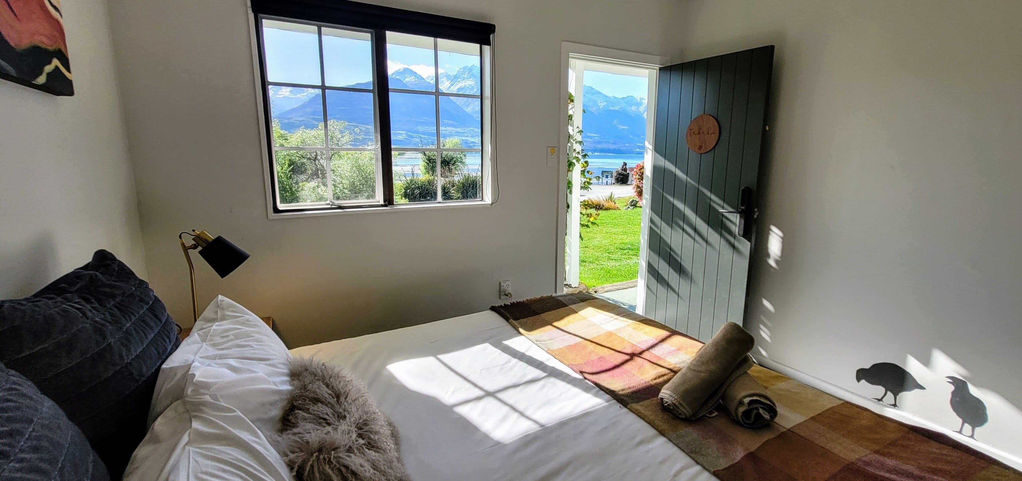 A bedroom with a large window, a bed with a fluffy pillow, a checkered blanket, and a view of mountains and a lake.
