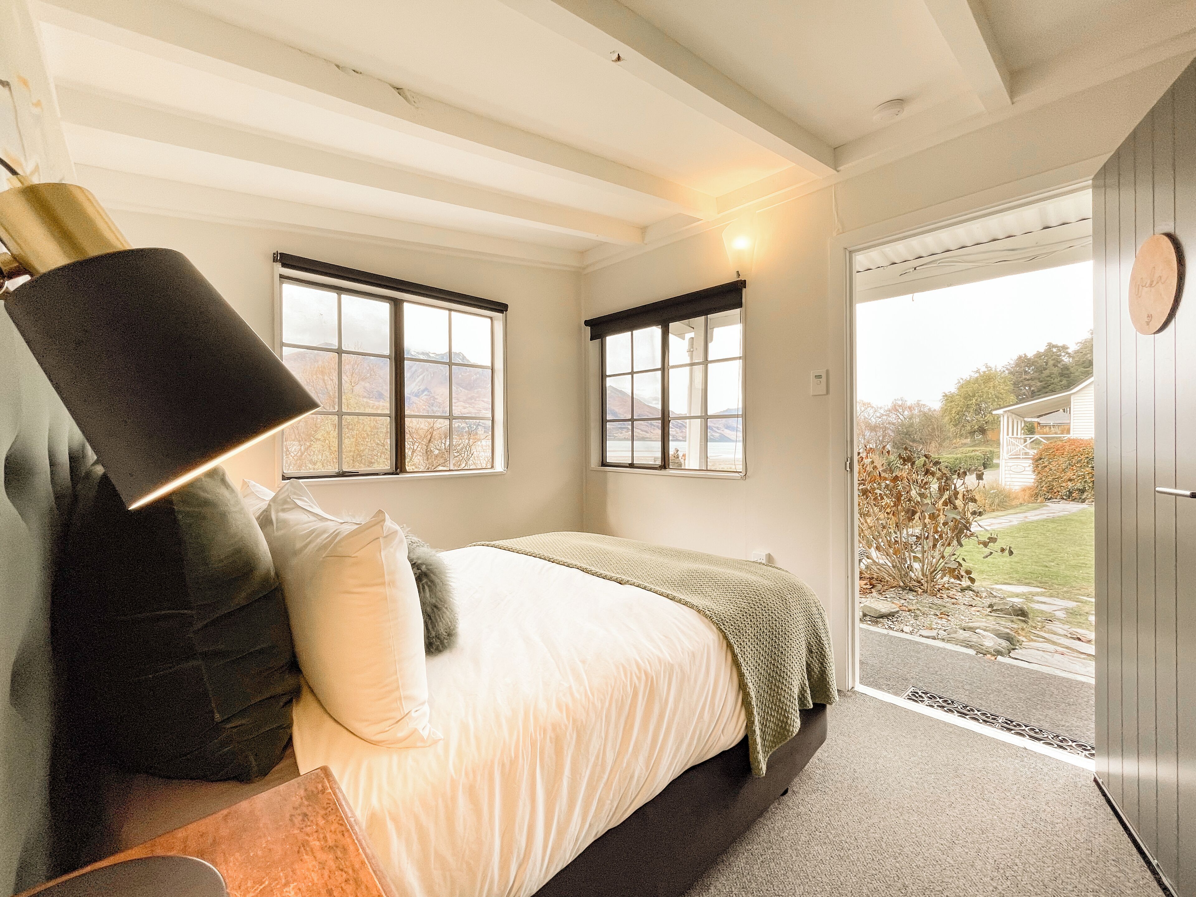 A bedroom with a bed, two windows, a lamp, and a view of the outdoors.