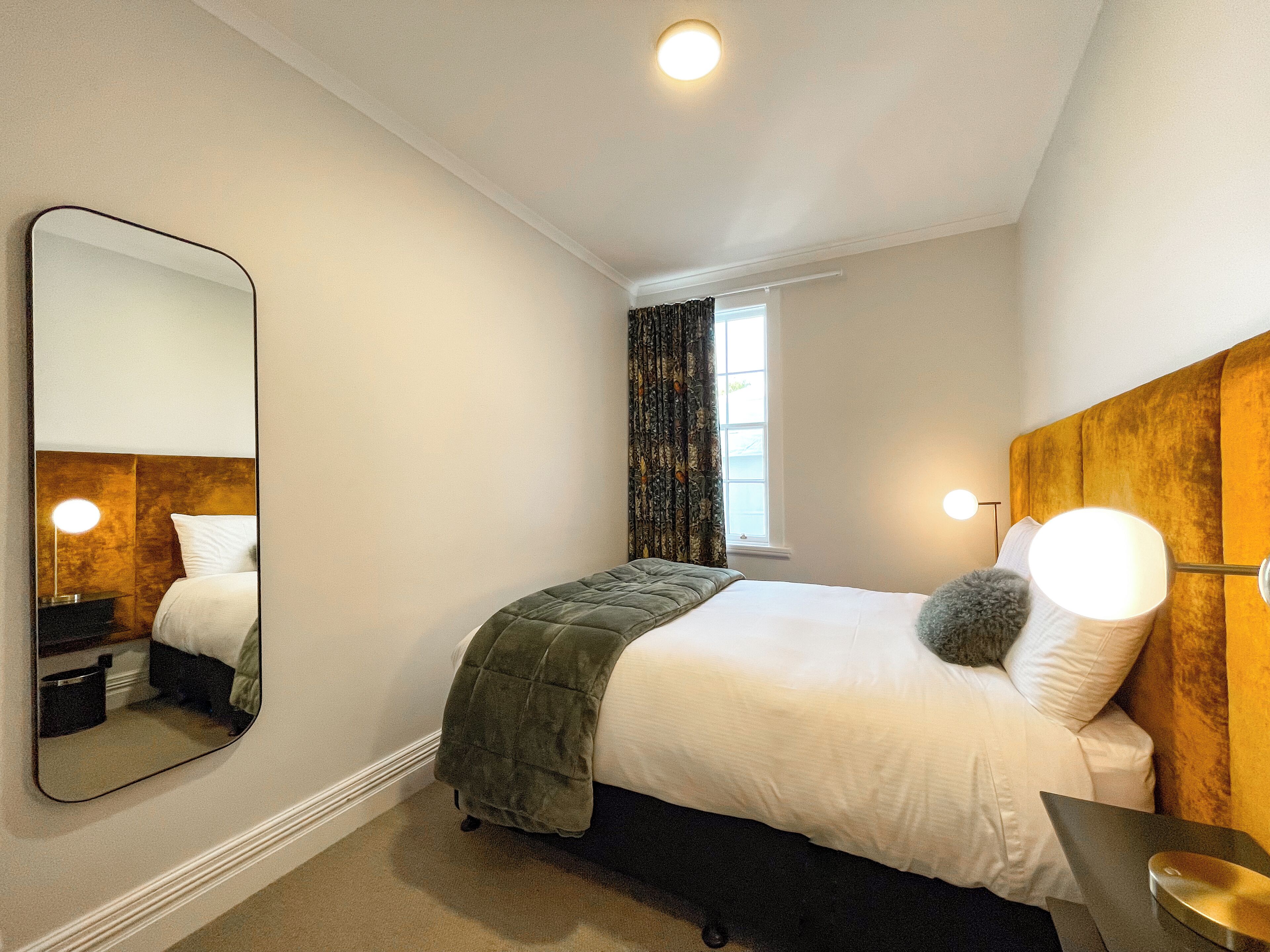 Un dormitorio con una cama, un espejo grande, una ventana con cortinas y una lámpara de noche.