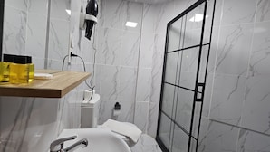 A modern bathroom with a marble backsplash, a wooden vanity, a white sink, a handheld hair dryer, and a glass shower enclosure.