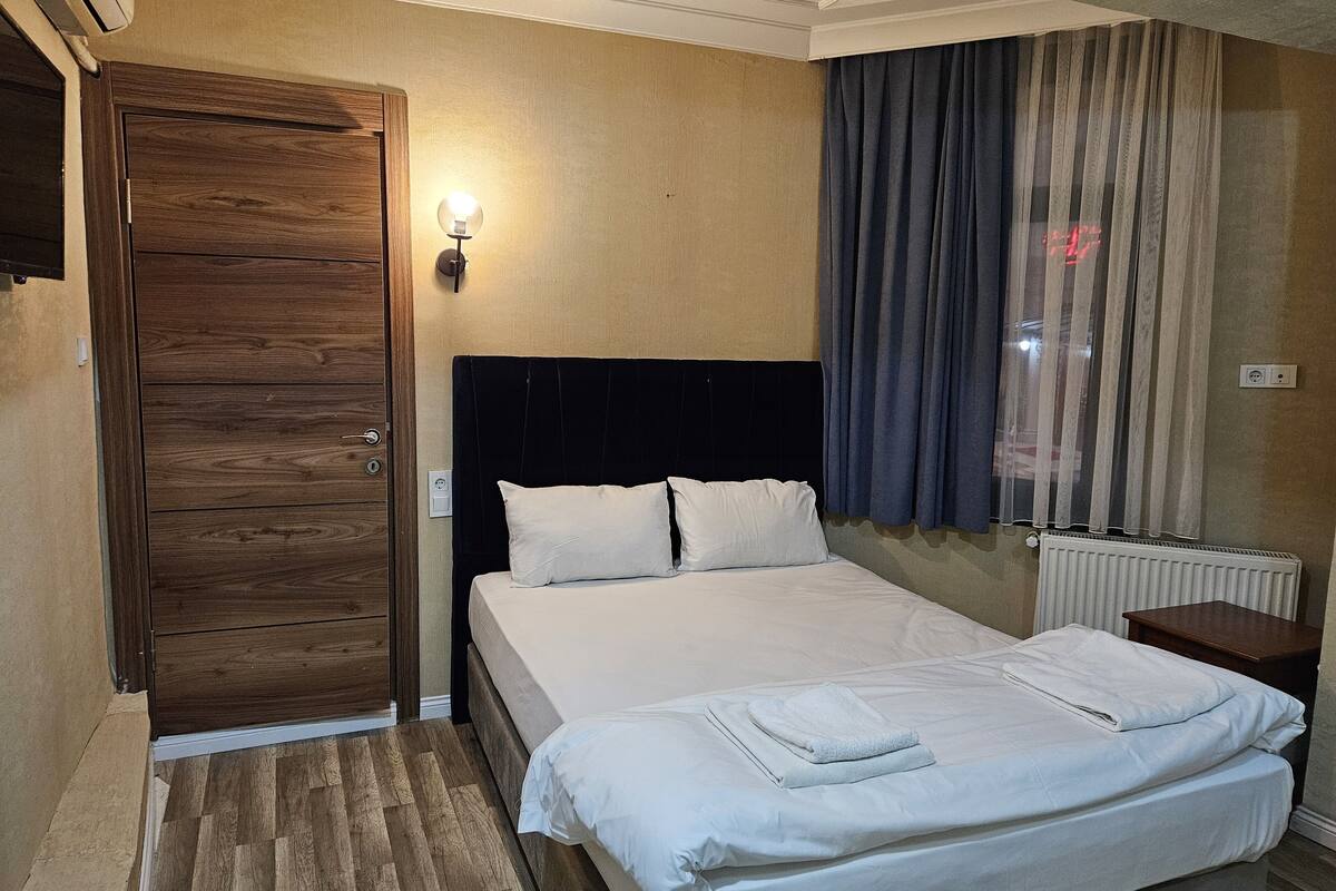 A hotel room with a wooden door, a bed with white linens, a bedside table, and a window with curtains.
