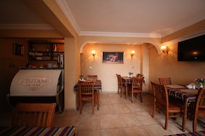 A hotel dining area with wooden tables and chairs, a buffet counter, wall-mounted lamps, and a flat-screen TV.