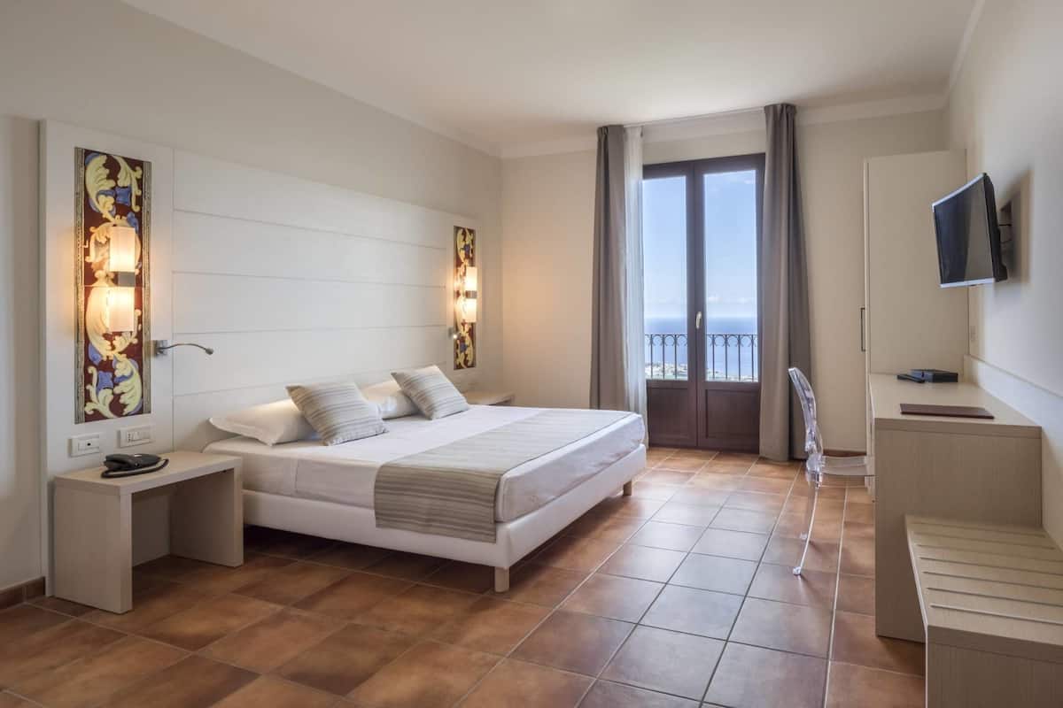 A bedroom with a bed, bedside tables, a TV, and a balcony with a view.