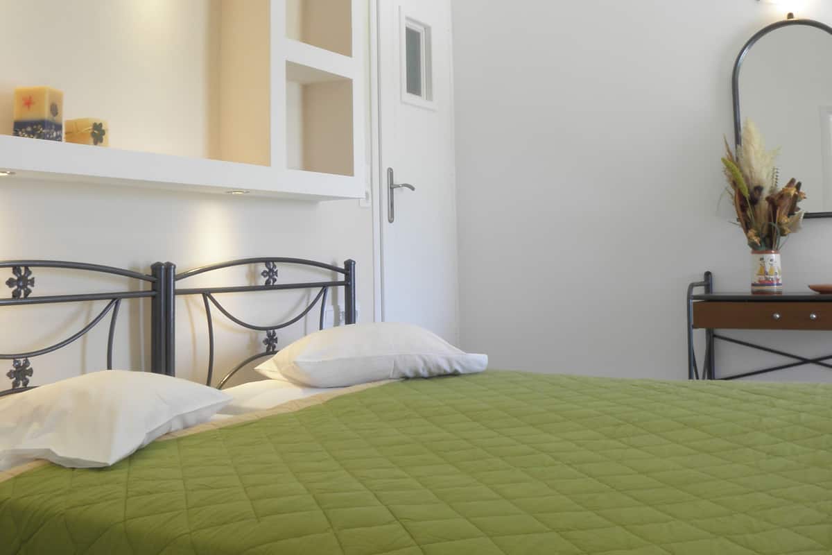 Um quarto com colcha verde acolchoada, uma porta branca com pequena janela, mesa de cabeceira com espelho e prateleira com objetos decorativos.