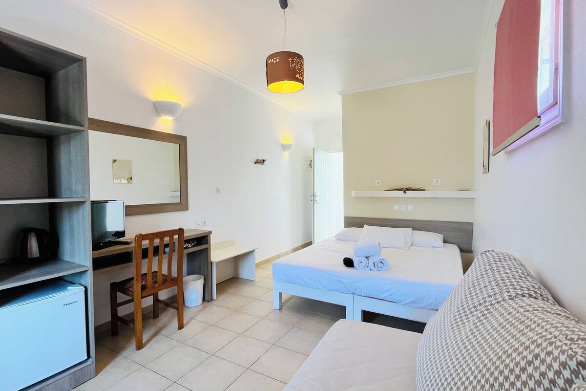 Quarto de hotel com cama, escrivaninha, cadeira, televisão e pequena cozinha.