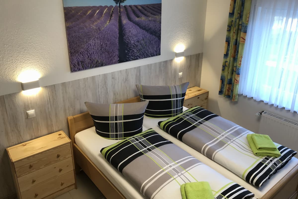 Un dormitorio con dos camas, mesitas de noche de madera, una lámpara de pared y un cuadro de un árbol en un campo.