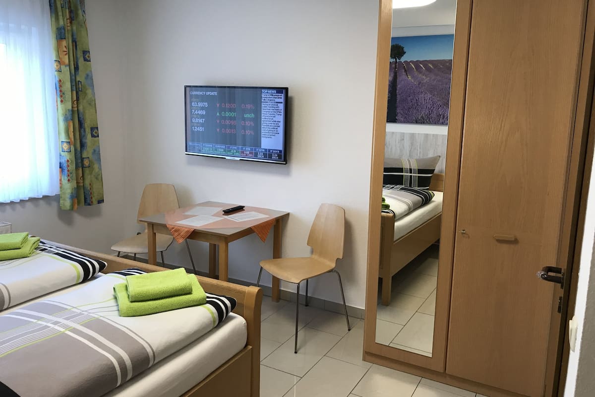 Habitación de hotel con dos camas, un escritorio, una silla, un televisor que muestra actualizaciones de divisas y un espejo que refleja la habitación.
