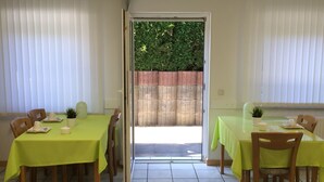 Comedor con mesas cubiertas con manteles verdes, sillas de madera y vista a una pared de ladrillo y follaje exterior.
