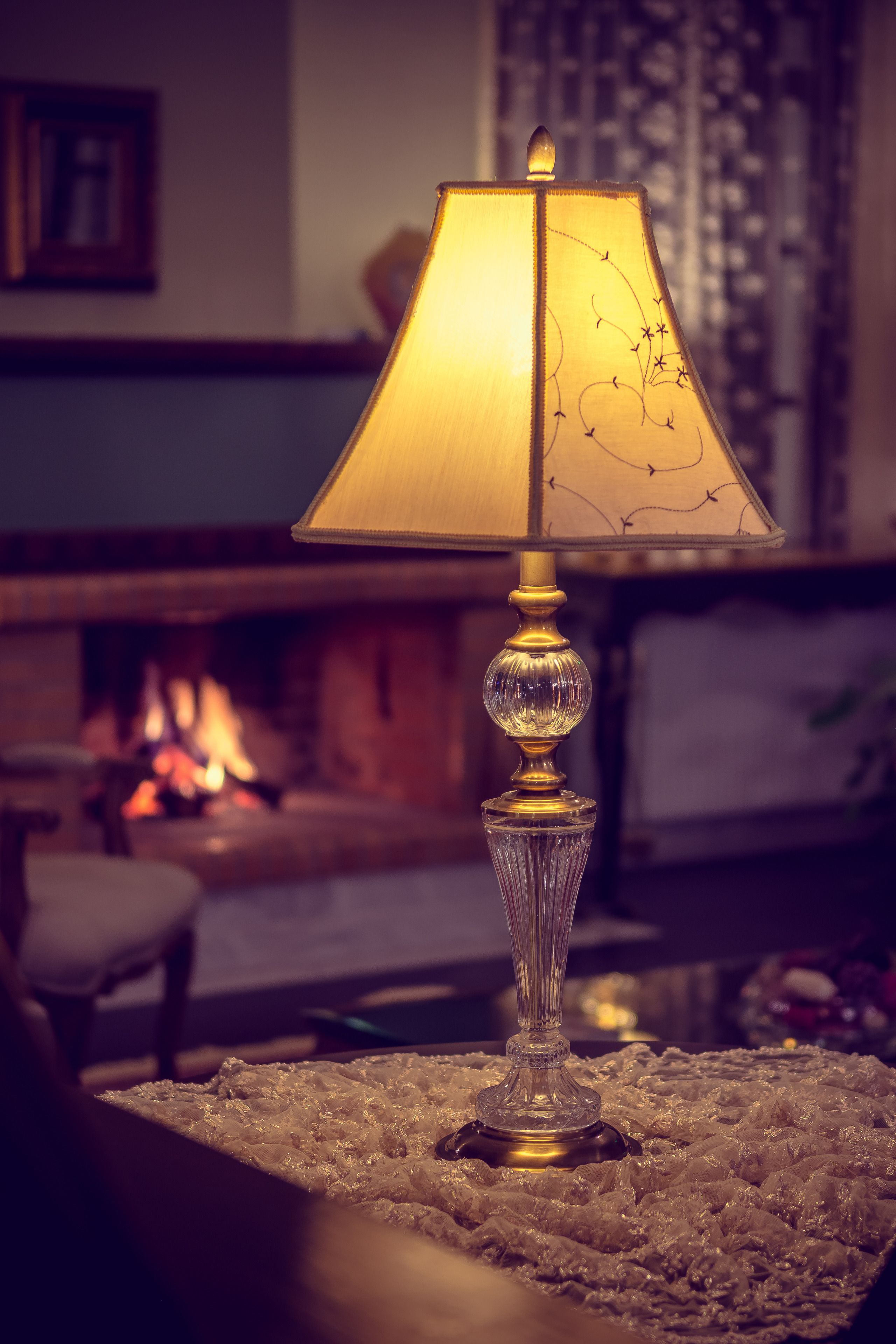 Una lampada da tavolo con base in vetro trasparente e paralume decorativo, posata su un tavolo dalla superficie strutturata in una stanza illuminata in modo caldo con un caminetto sullo sfondo.