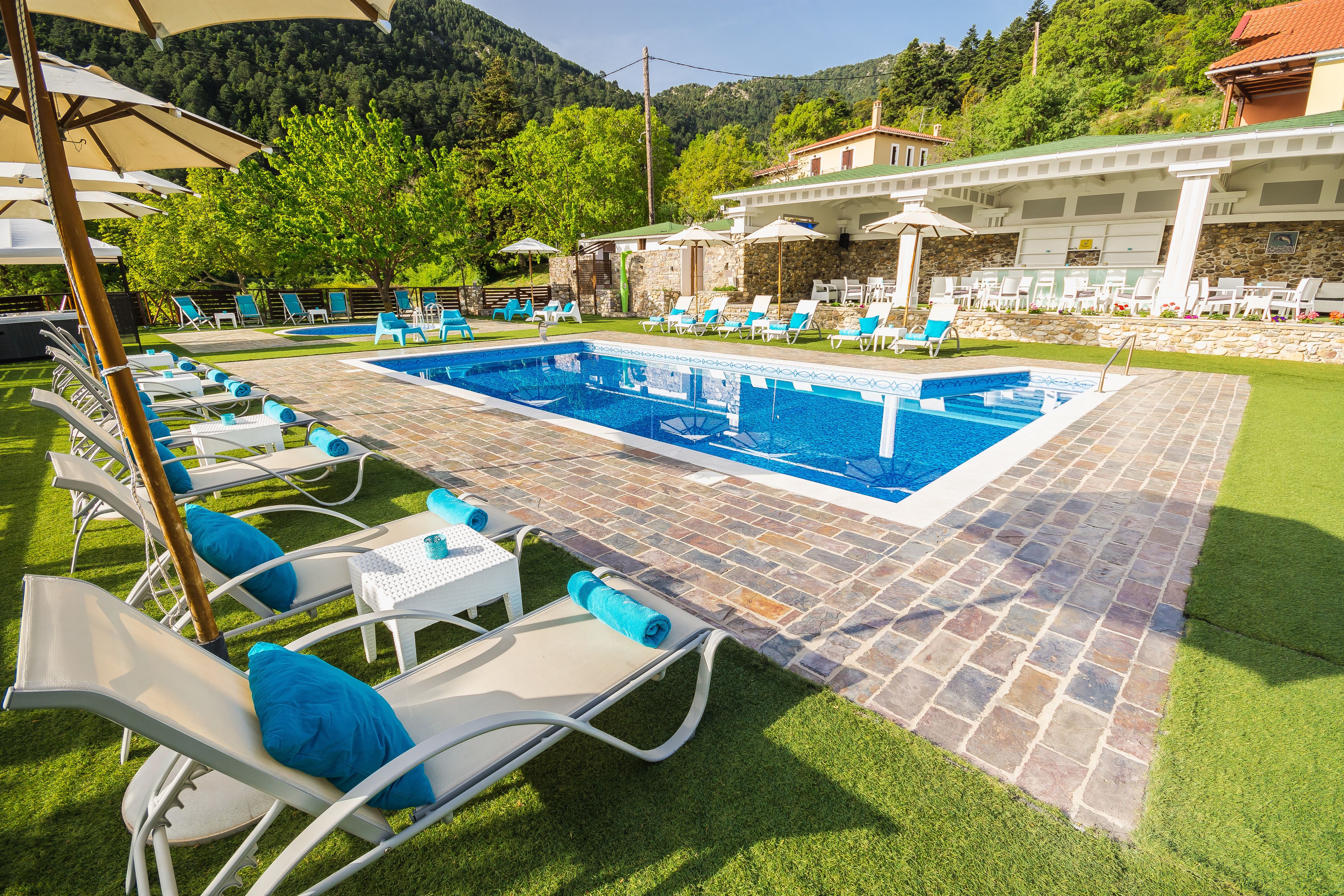 Ein Poolbereich mit Liegestühlen und Sonnenschirmen, umgeben von einer Steinterrasse und üppiger Bepflanzung.