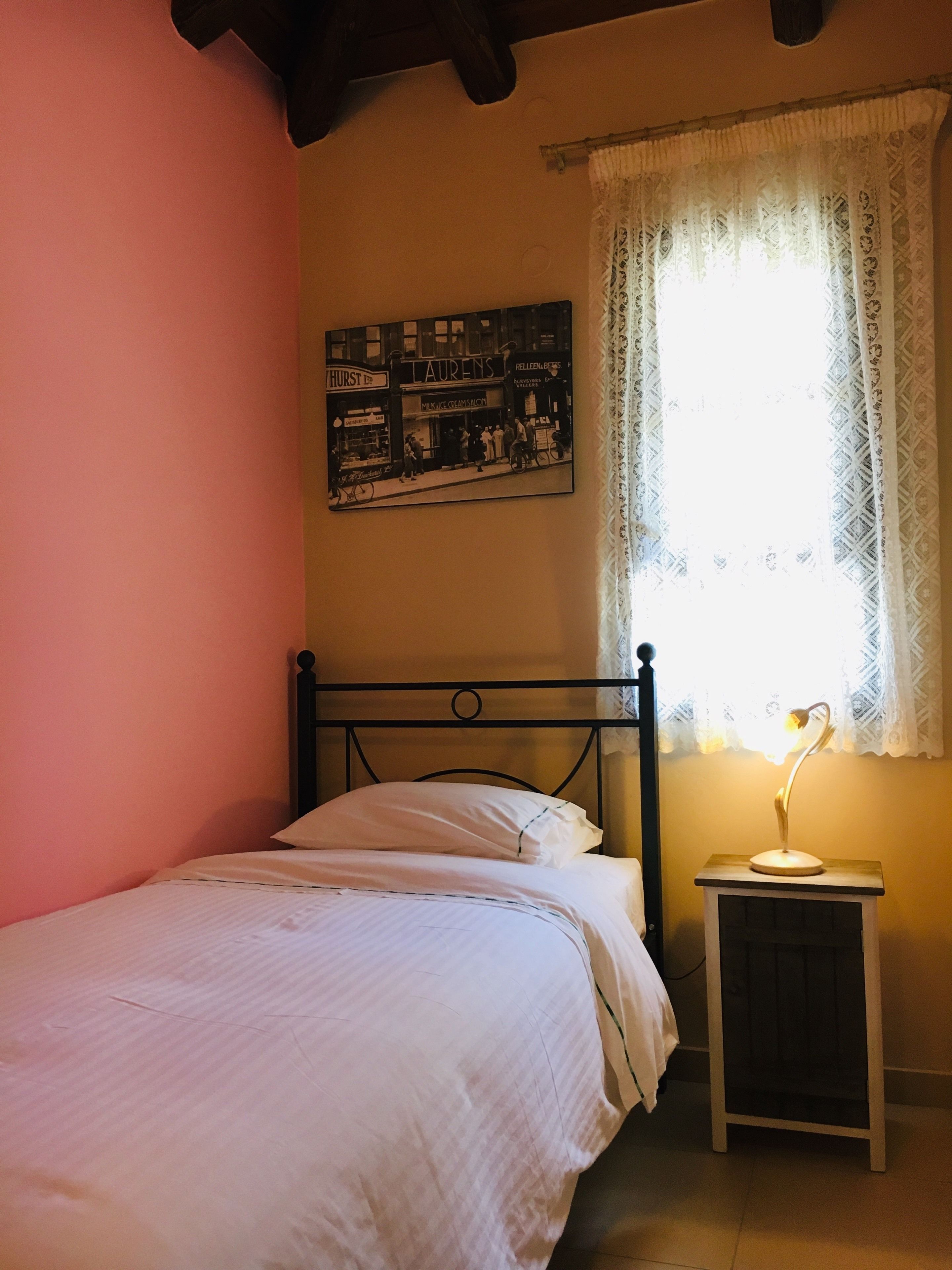 Une chambre à coucher avec un lit, une table de chevet avec une lampe, une fenêtre avec des rideaux en dentelle et un mur orné d’un tableau encadré.